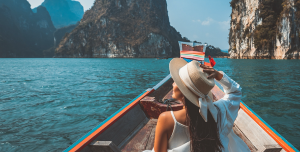 A woman sits in a boat in an exotic location, holding her hat.