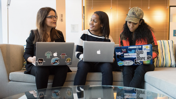 Three women seated on a sofa having a conversation over their laptops